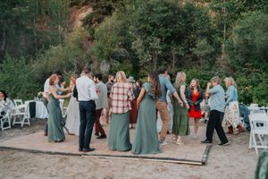 Wedding guests dance together on an outdoor dance floor surrounded by trees and white folding chairs.
