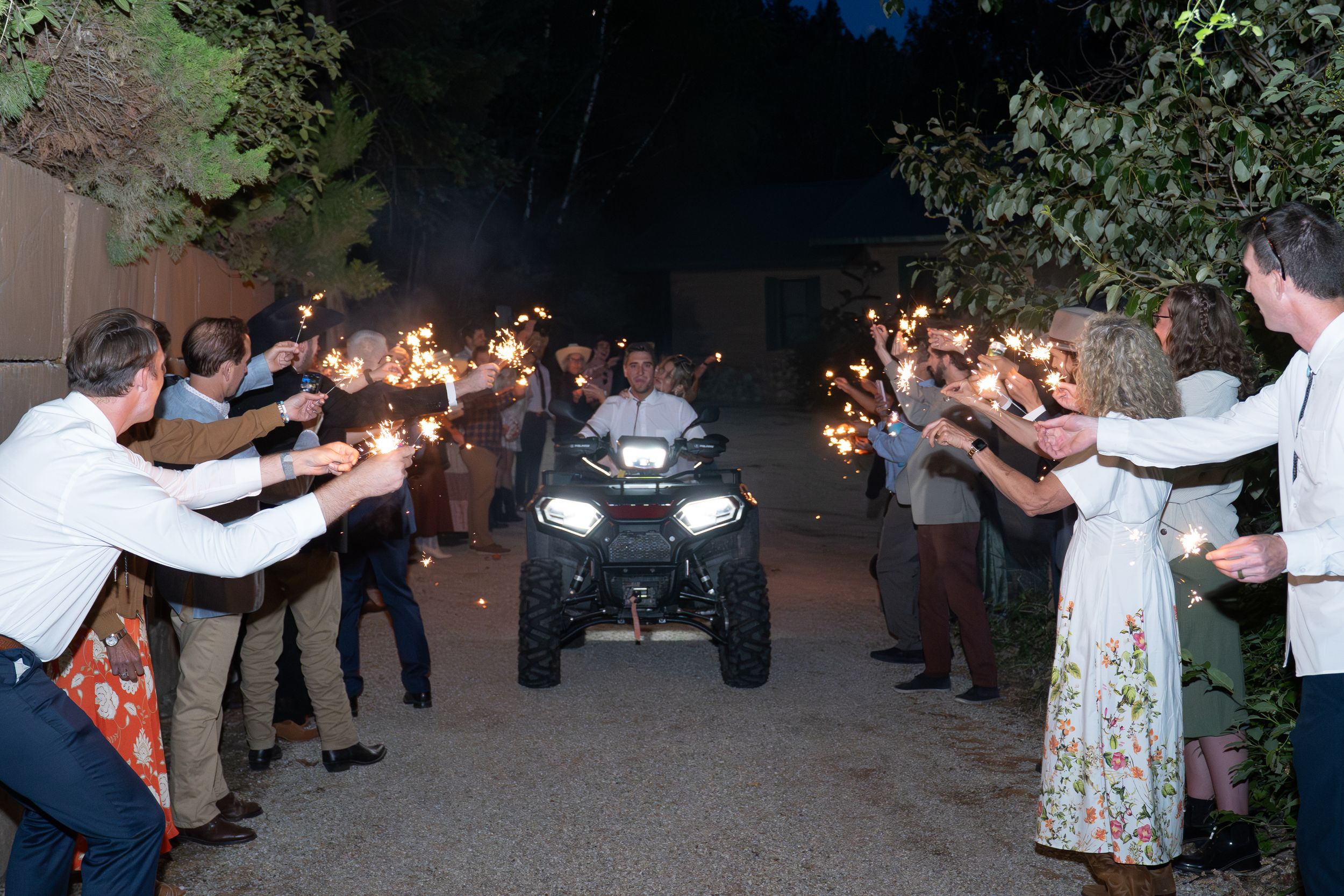Wedding guests and bridal party form a long human tunnel under twinkling lights at an outdoor evening reception.