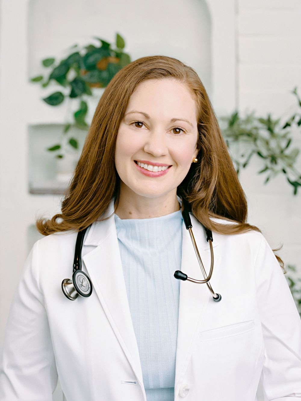 Healthcare professional in white coat and light blue sweater poses with stethoscope against bright white background with plants.