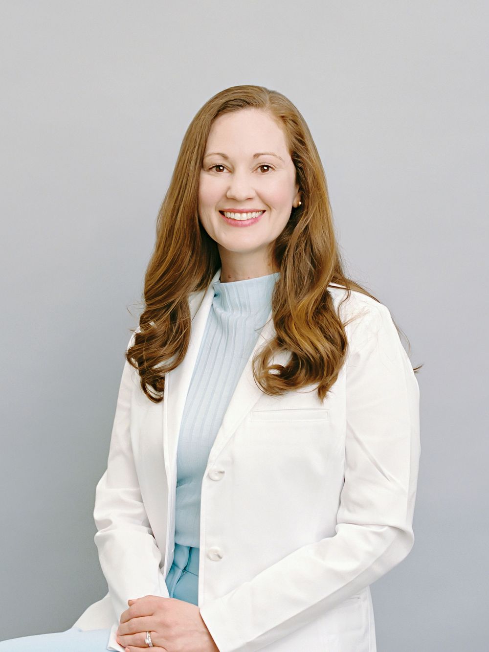 Professional portrait of someone in a white lab coat and light blue sweater against a neutral grey background.