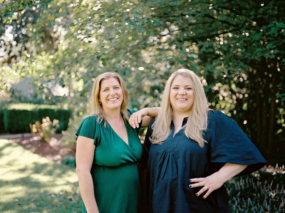 Two women in green and navy blue dresses pose together outdoors in a sunlit garden setting.