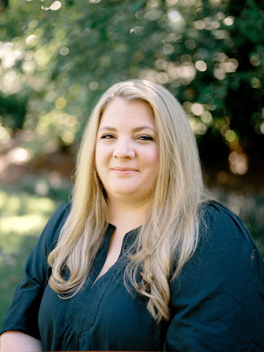 A person with blonde hair poses outdoors in a navy top against a softly blurred natural background of green foliage.