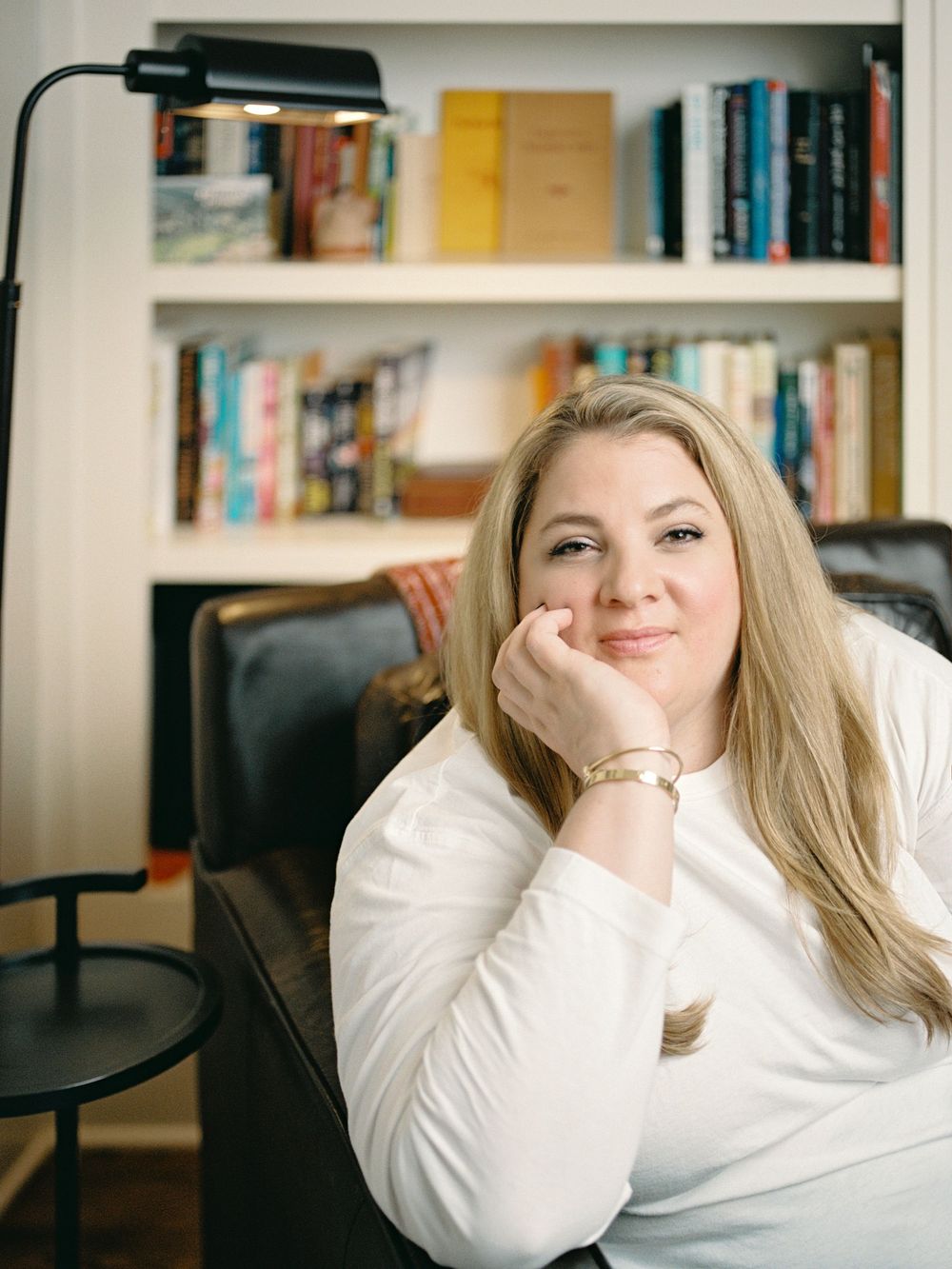 A person in a white sweater smiles warmly while resting their chin on their hand in front of a bookshelf.