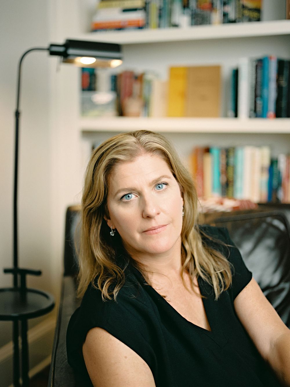 A person in a black top sits casually in front of bookshelves and a reading lamp in a home library setting.