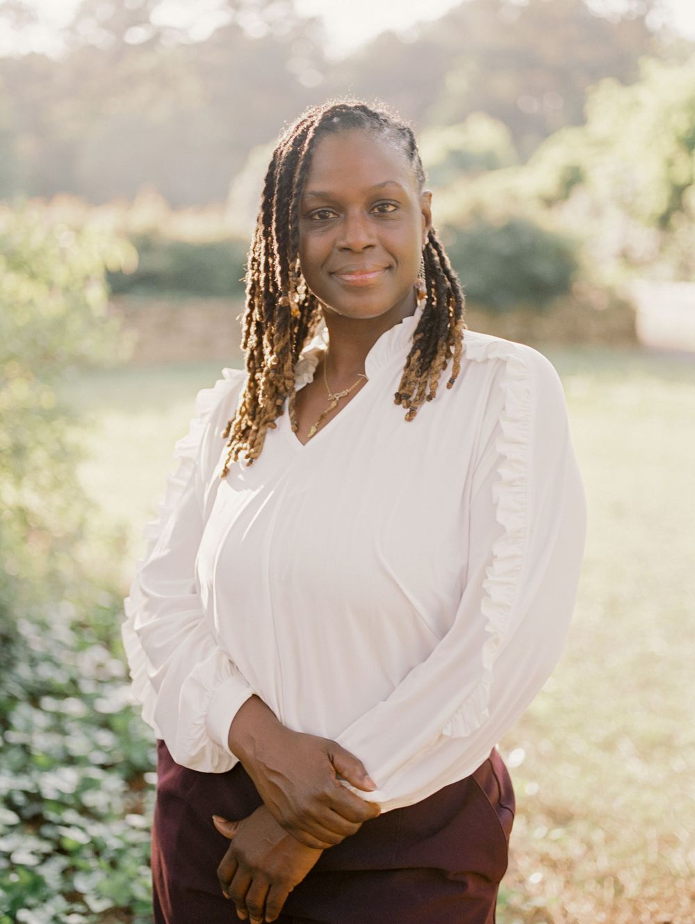 Person with braided hair wearing a white blouse and burgundy skirt stands outdoors in natural sunlit setting.