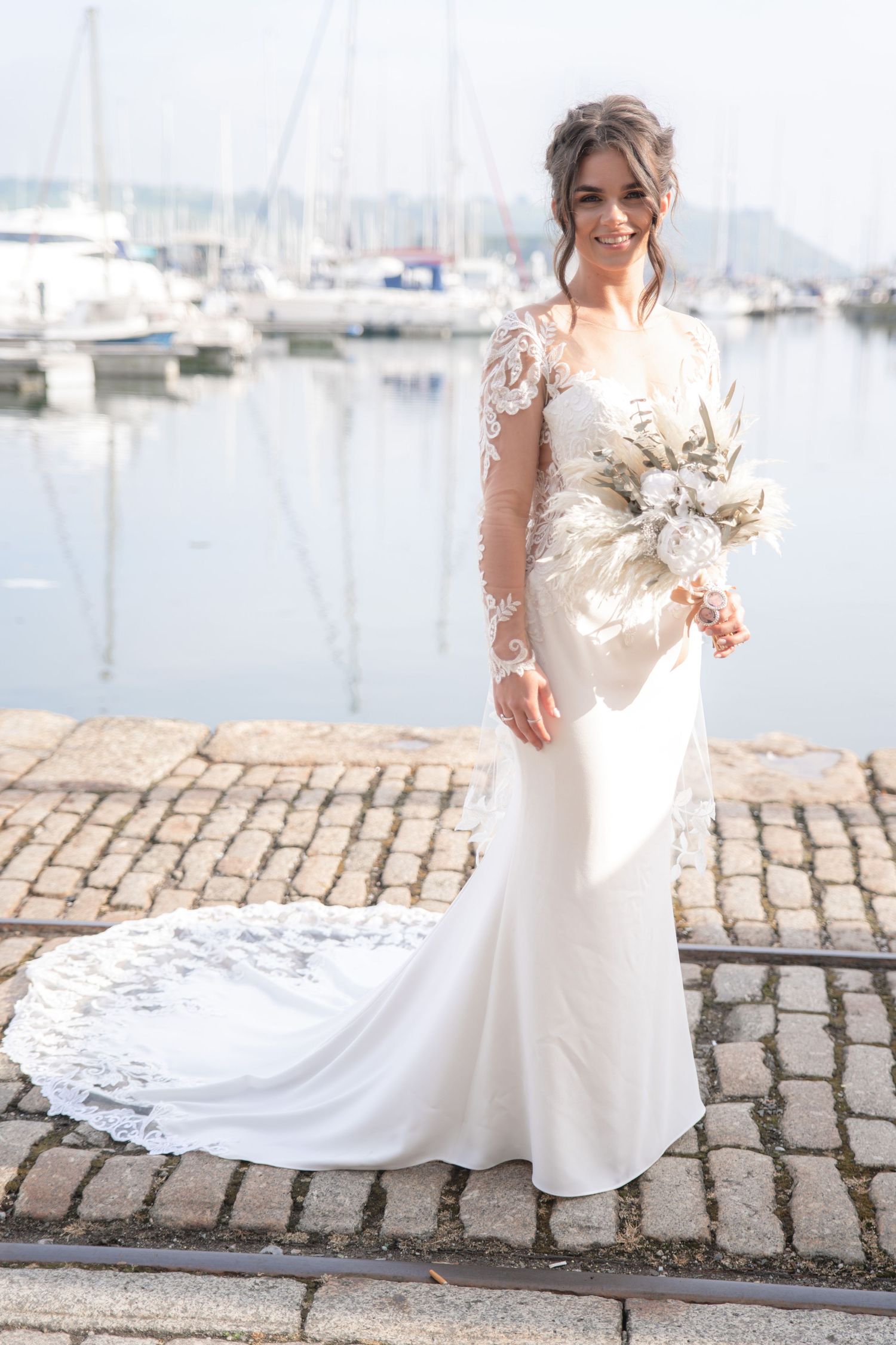 Elegant bride in white lace wedding dress standing on cobblestone walkway near marina on wedding day.