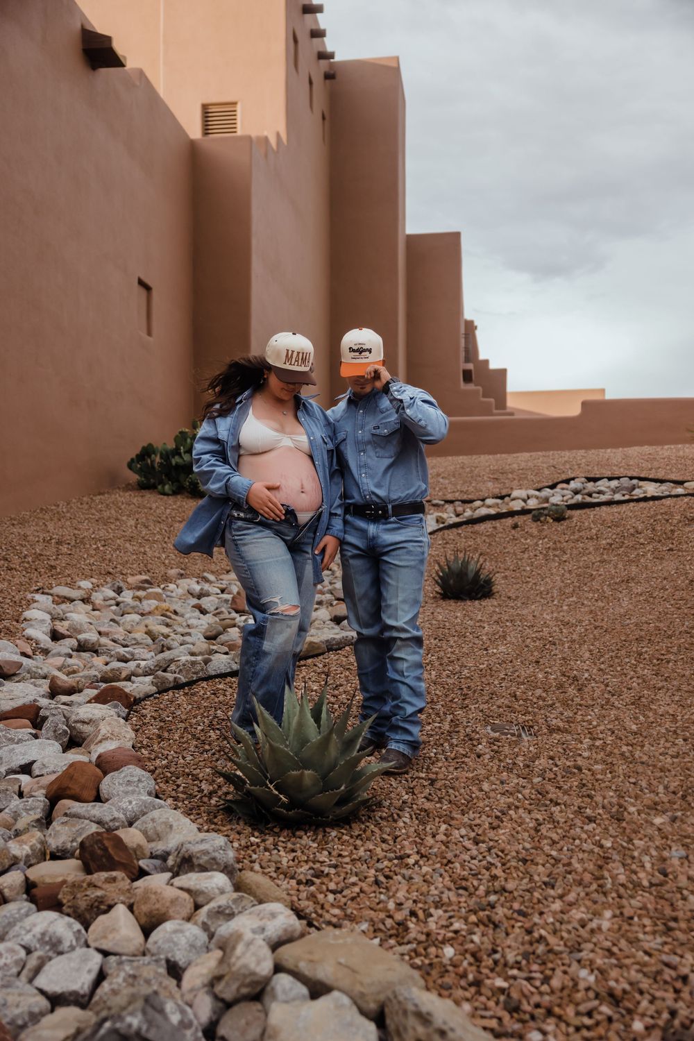Pregnant couple in matching denim outfits and trucker hats pose near adobe building with desert landscaping.