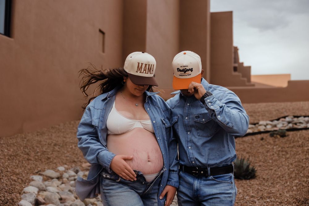 Pregnant woman wearing Mama trucker hat stands with partner in denim near adobe wall.