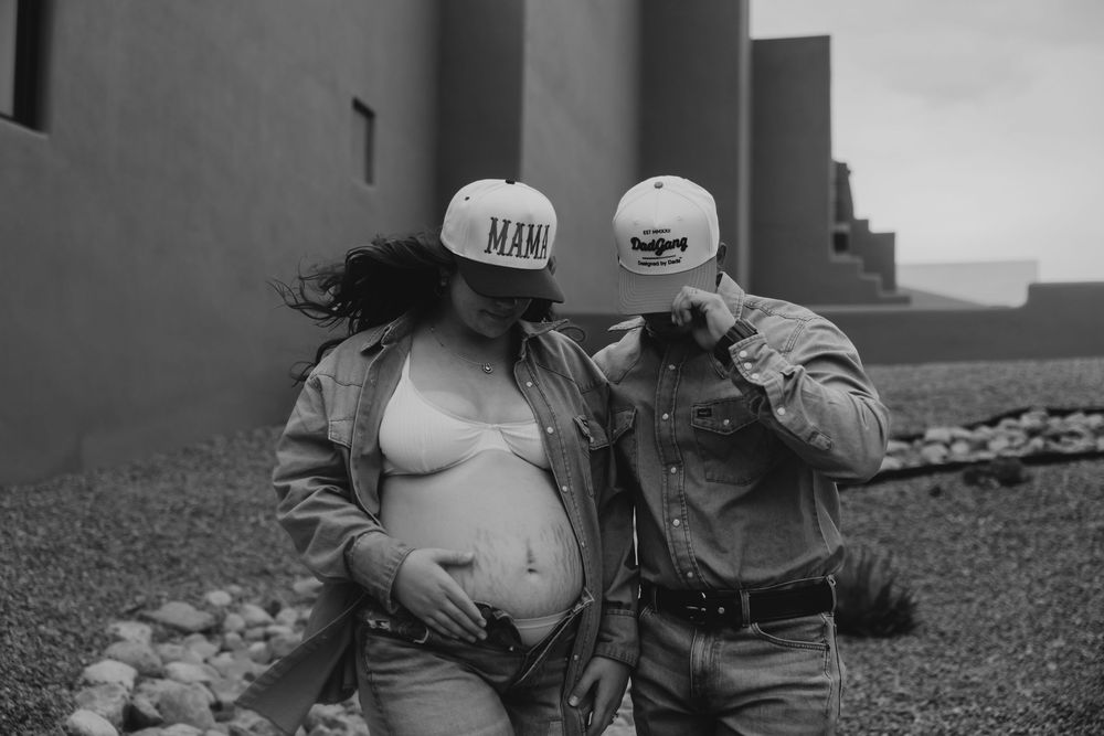 Black and white maternity photo of couple in trucker hats embracing near adobe building.