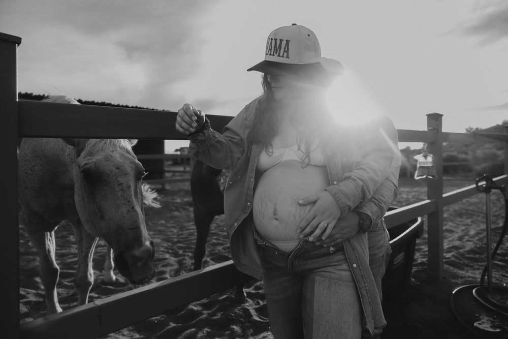 Black and white photo of pregnant woman cradling baby bump near horses at golden hour.