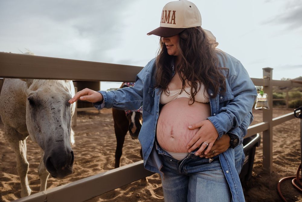 Pregnant woman in Mama cap gently pets white horse while holding baby bump at ranch.