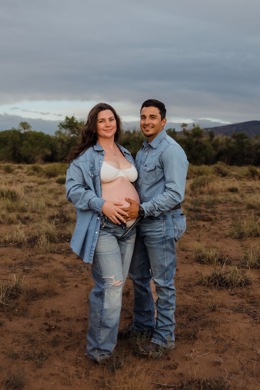 Expecting couple in matching denim embracing in desert landscape at dusk.