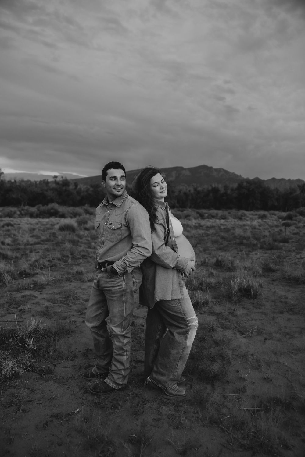 Black and white maternity photo of couple standing back to back in open field.