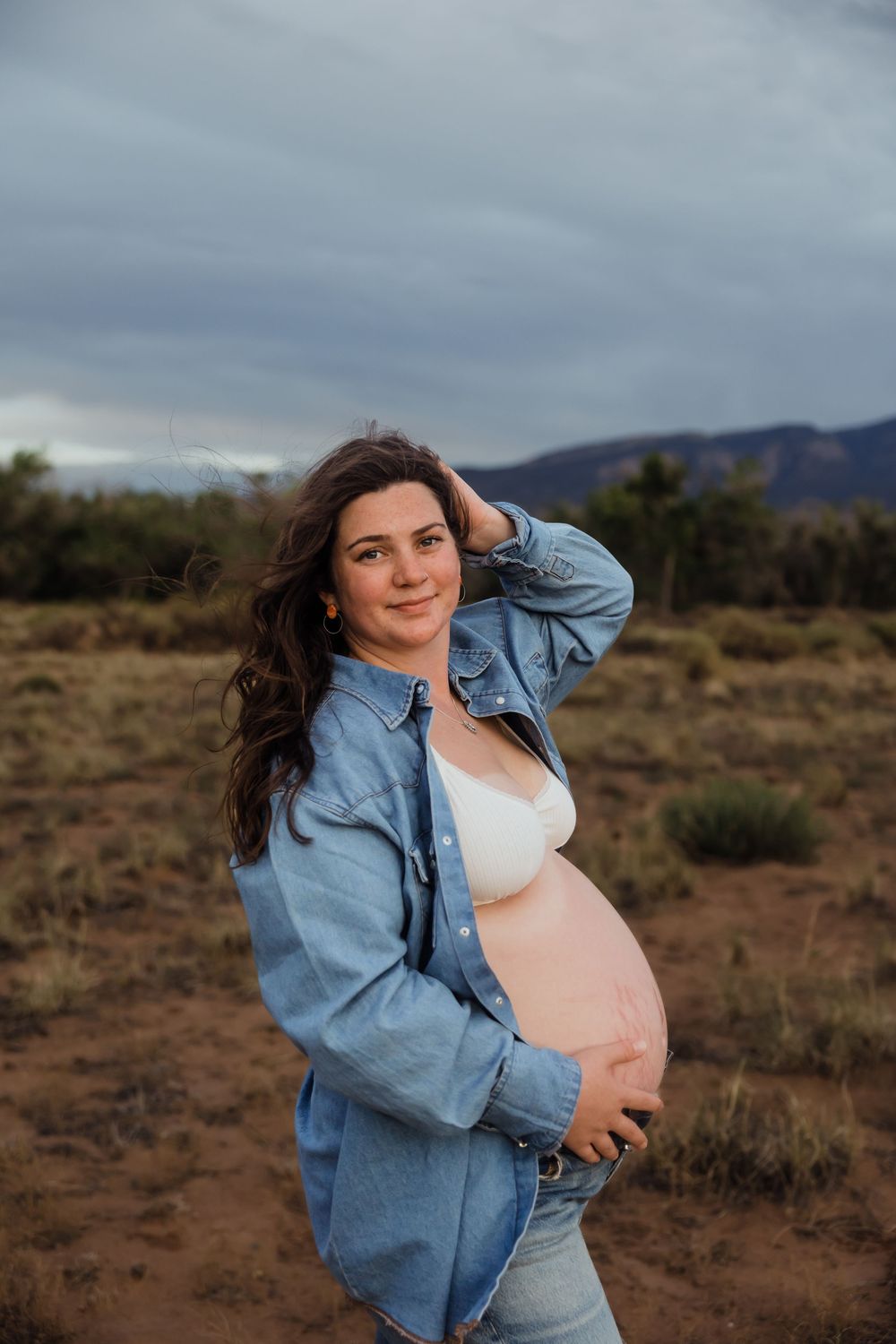 Smiling pregnant woman in denim jacket cradling baby bump in desert landscape.