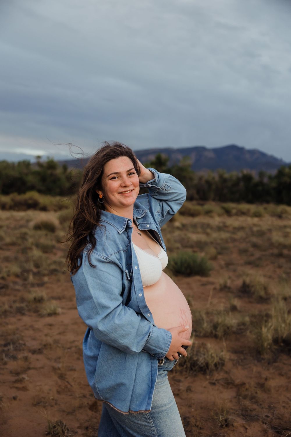 Glowing pregnant woman in denim jacket holding baby bump in scenic desert field.