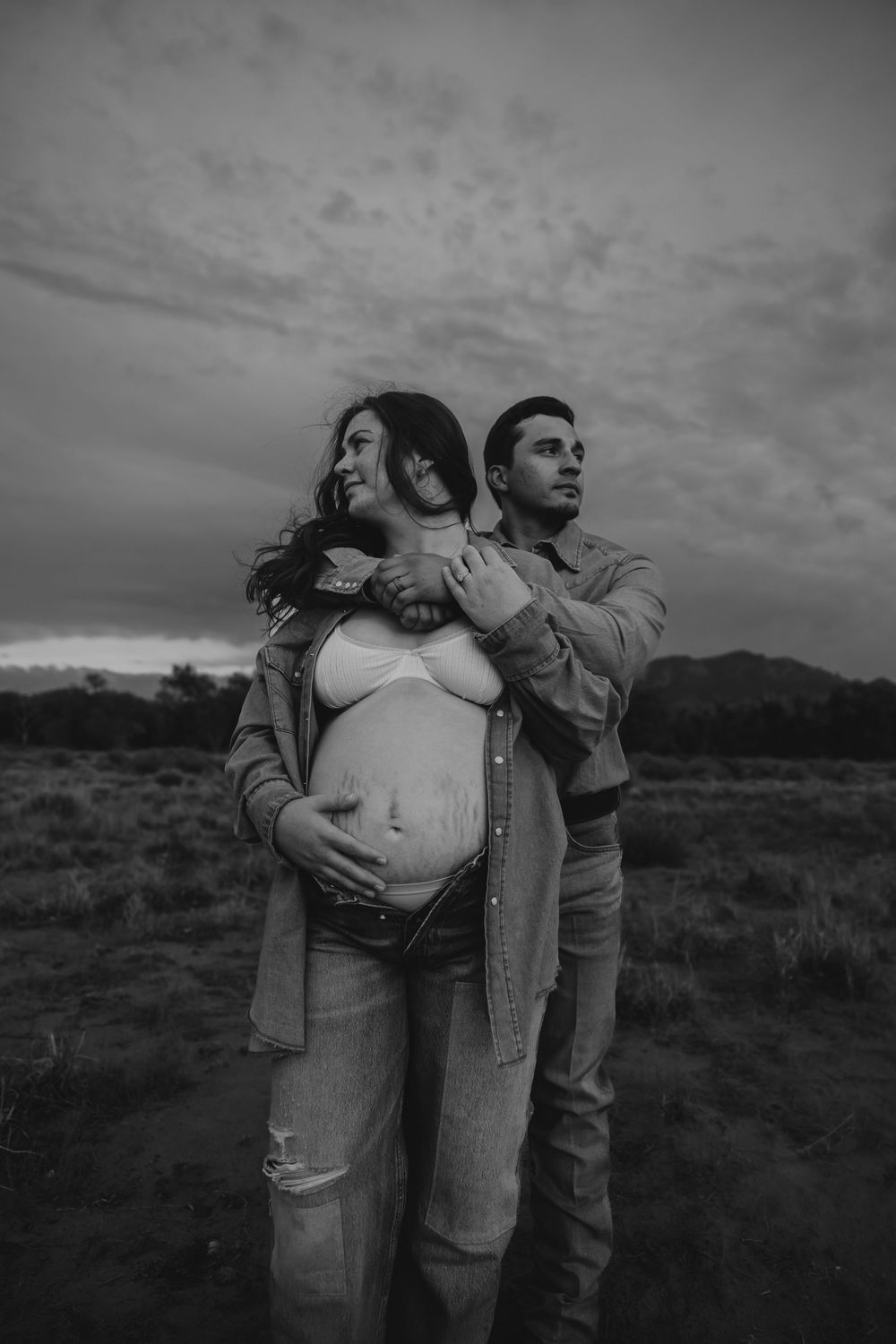 Black and white maternity photo of couple embracing in dramatic cloudy field setting.