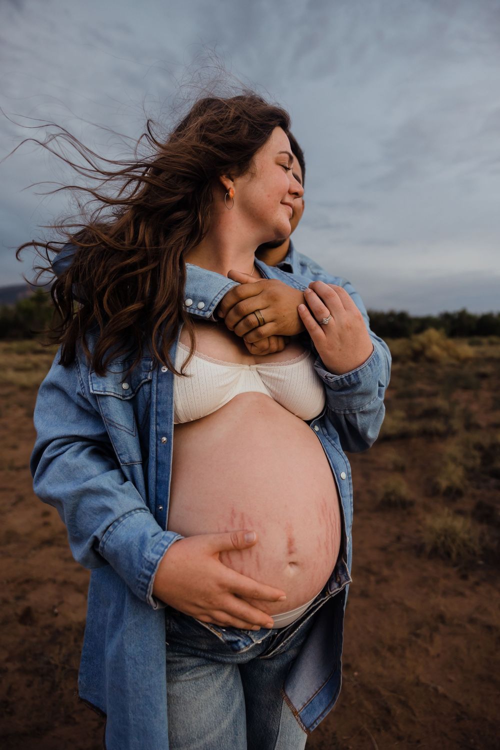 Pregnant woman in denim jacket holds baby bump outdoors with wind-blown hair against cloudy sky.