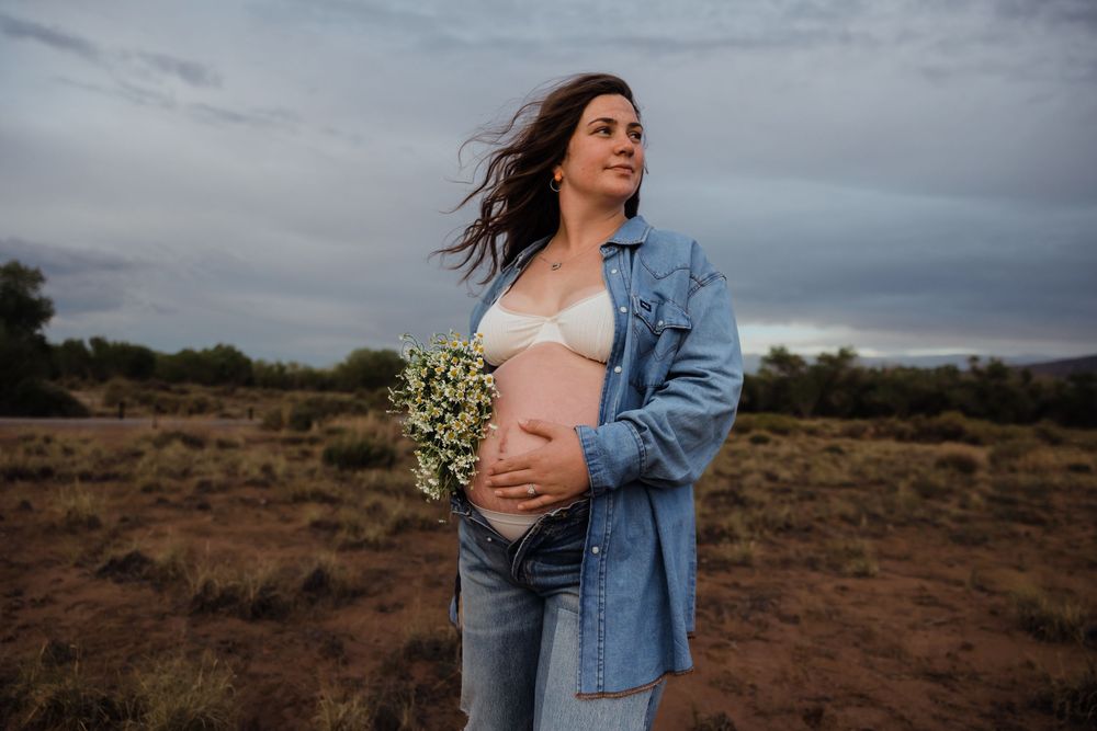Pregnant woman in denim shirt holds wildflowers and baby bump in open desert landscape.