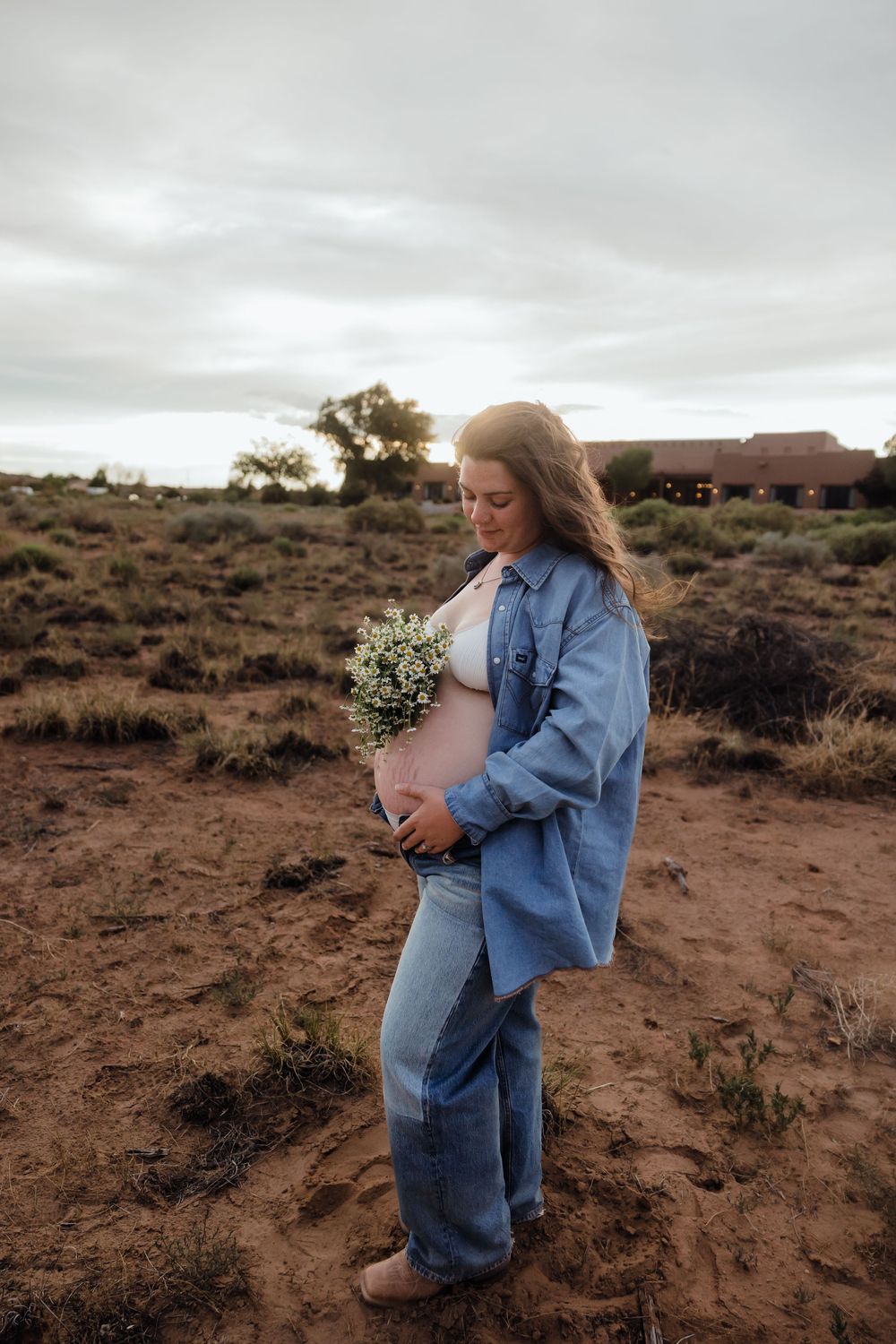 Pregnant woman in denim jacket cradles baby bump holding white wildflowers in sandy desert setting.