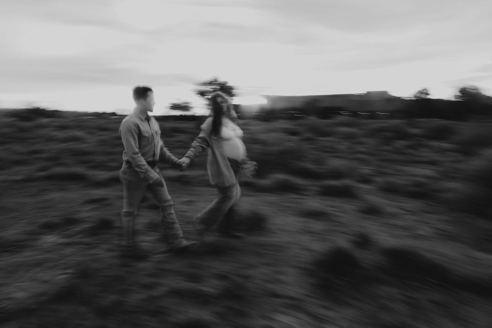 Black and white photo of couple holding hands walking through desert field during maternity shoot.