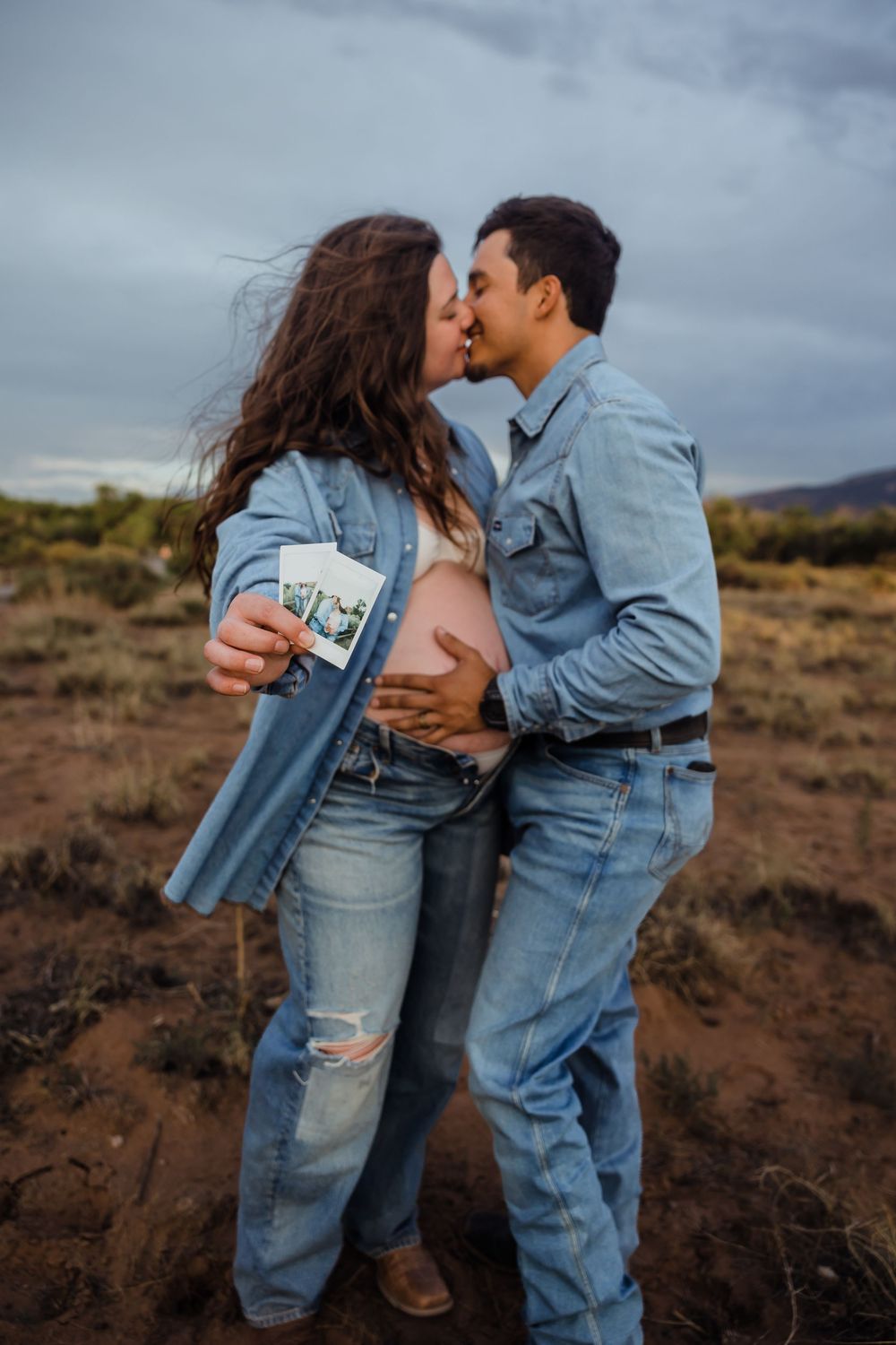 Couple kissing during maternity shoot outdoors, woman holding ultrasound photos while partner cradles bump.
