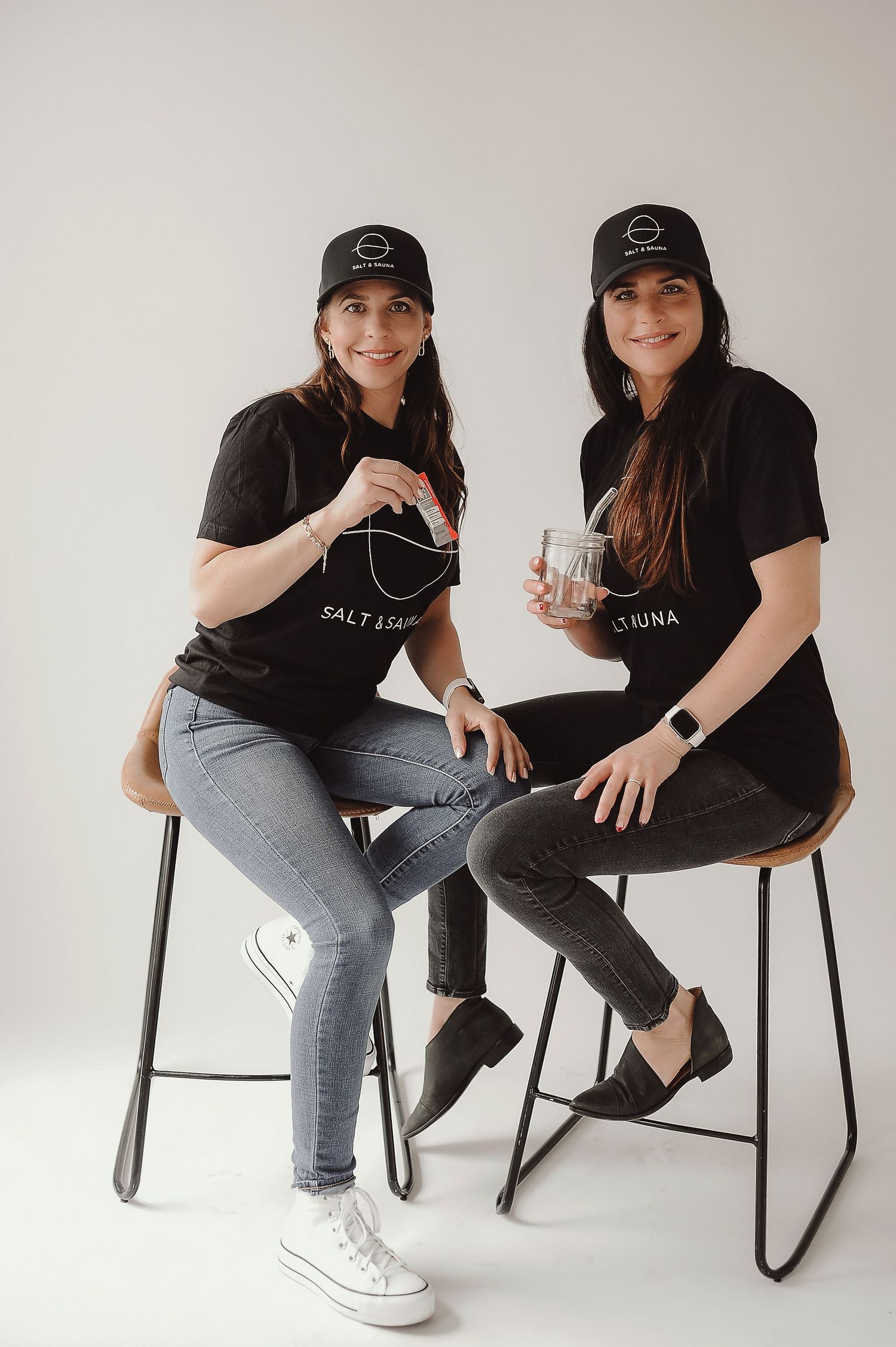 Two people in black shirts and jeans sitting casually on modern chairs against white background.