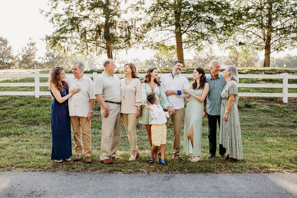 A happy wedding party poses together outdoors wearing coordinated navy and beige attire with white fencing in background.