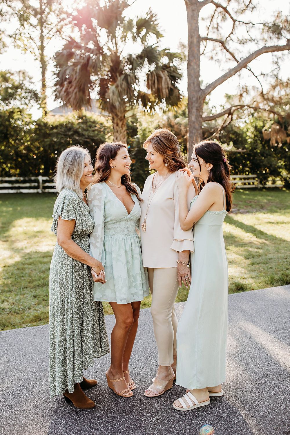 Group wearing light pastel dresses sharing joyful moments together on a paved path at sunset.