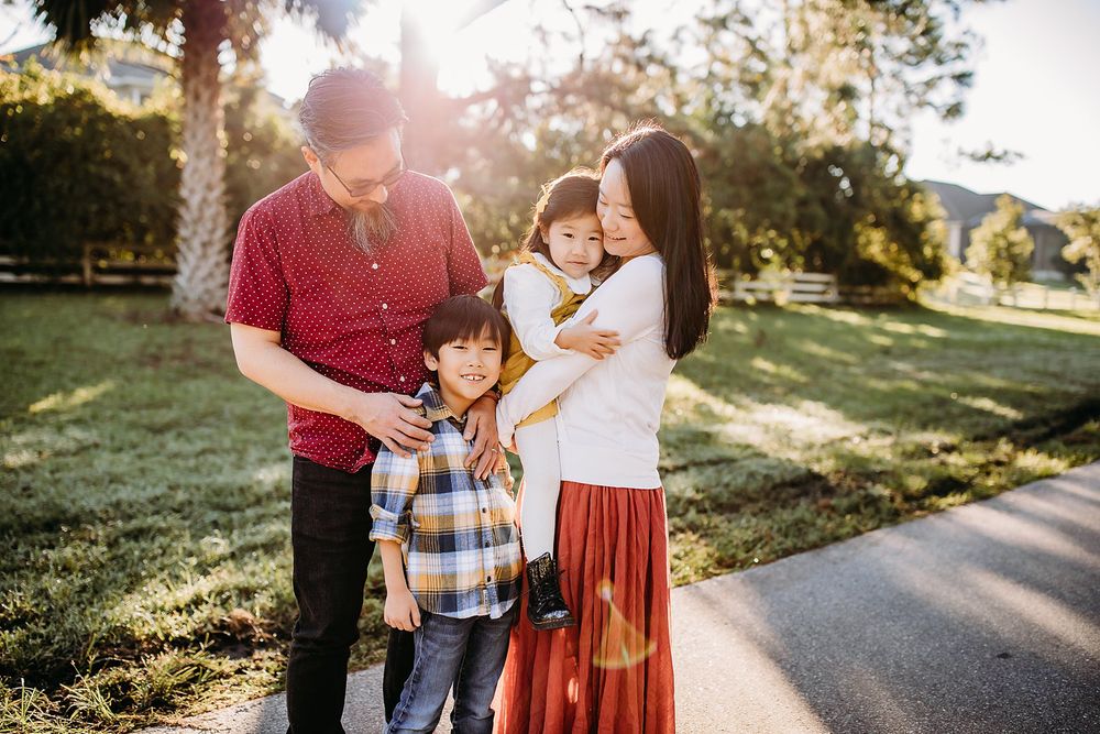 A family poses together outdoors on a path surrounded by sunlit trees.