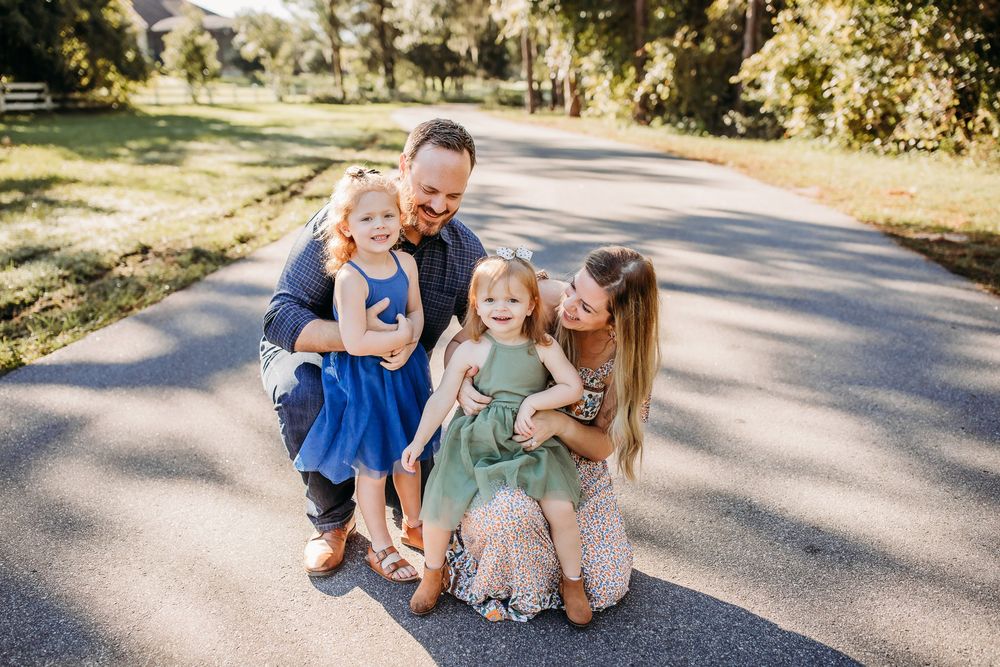 Family members embrace and laugh together while sitting on a sunlit pathway surrounded by trees.