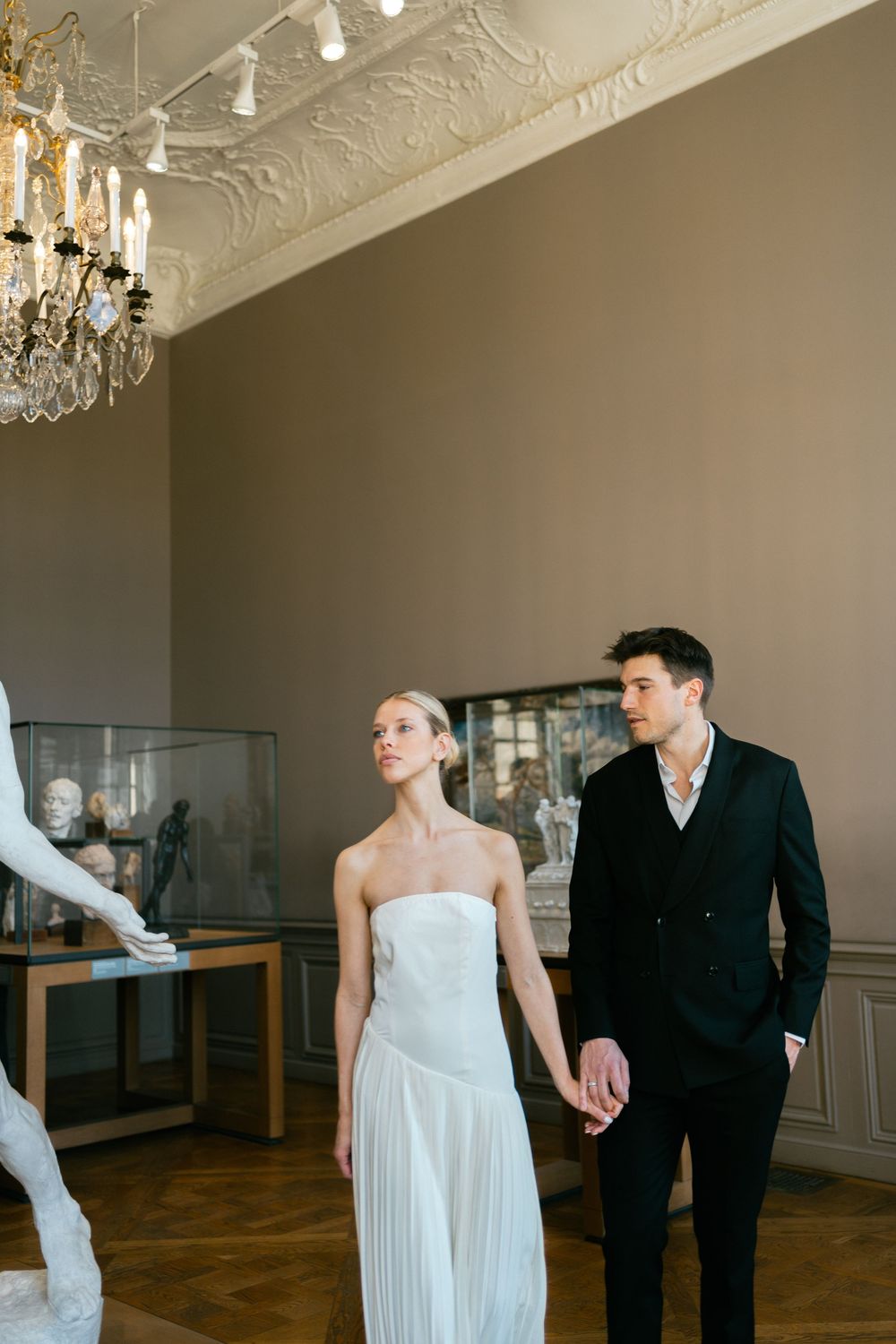 Couple in formal wedding attire holds hands while walking through an elegant room with ornate ceiling and chandeliers.