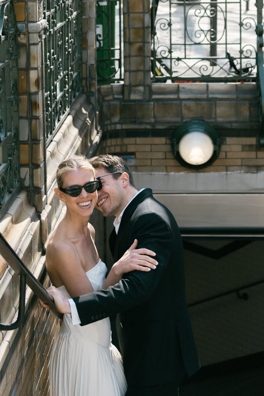 A joyful couple share a tender moment on a sunny day against a brick building with ornate ironwork.