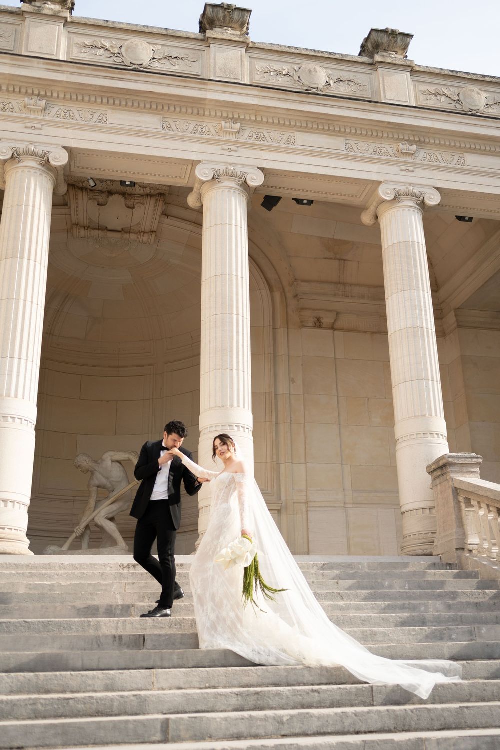 A couple in wedding attire sits on classic stone steps with white ionic columns in an elegant architectural setting.