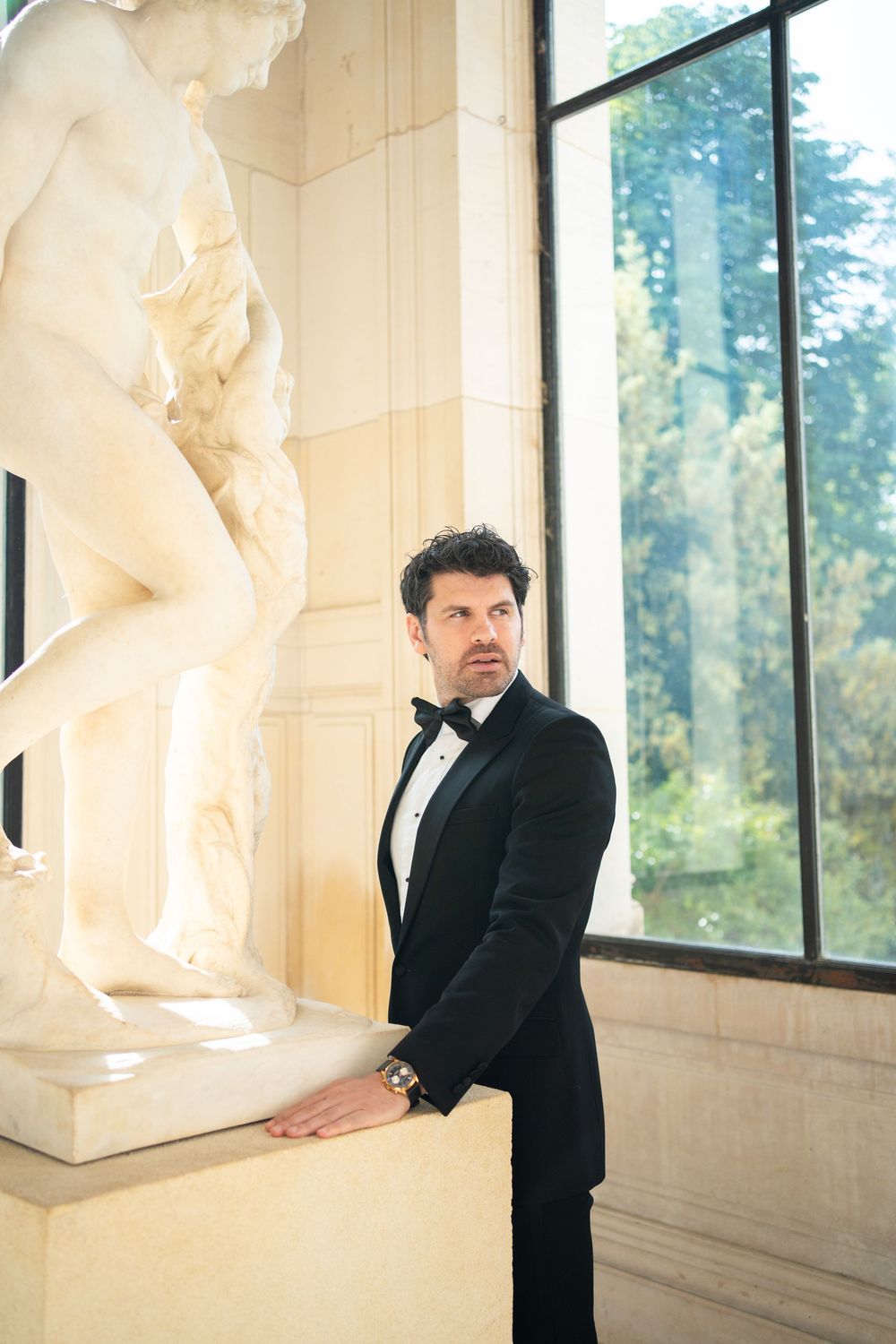 A person in a black tuxedo stands next to a classical marble statue near a window in an elegant interior setting.