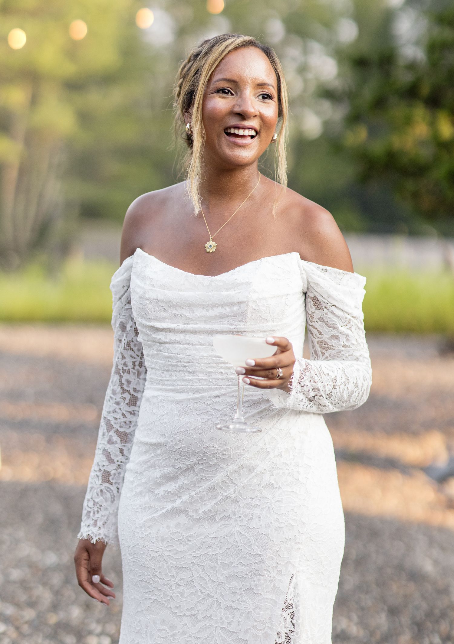 Laughing, joyful woman in flowing white lace dress twirls playfully outdoors in golden afternoon sunlight.