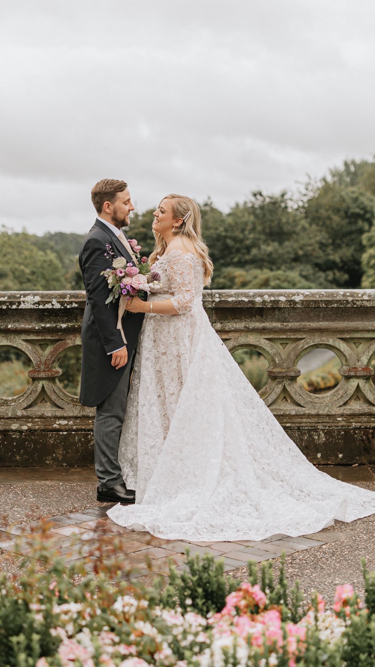 A bride and groom share a romantic moment on a stone terrace surrounded by pink garden flowers in bloom.