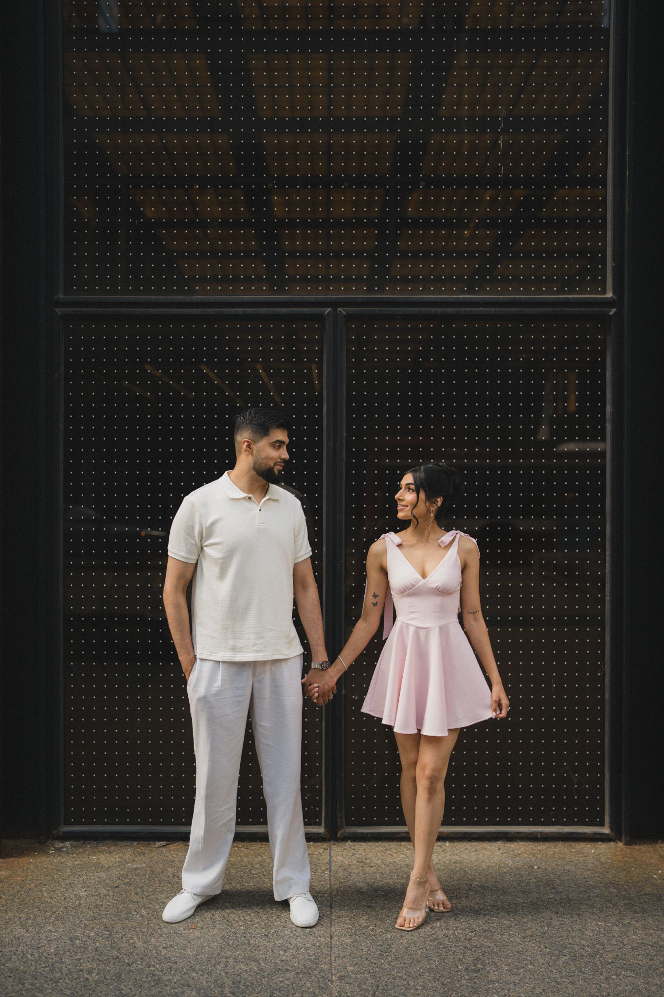 Couple in coordinated white and pink outfits holding hands while posing against an industrial gray wall.