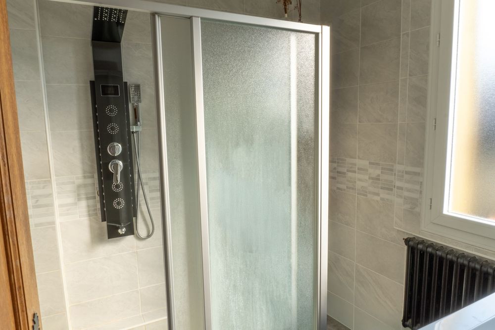 Modern bathroom interior with frosted glass shower door and black shower panel mounted on gray tiled wall.