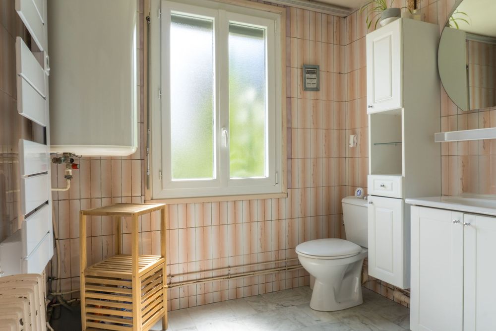 Pink striped bathroom interior with white fixtures, wooden storage shelf and natural light from window.