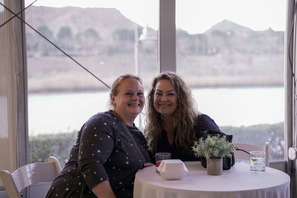 Two people sit at a small round table with coffee cups and flowers against a scenic mountain backdrop near water.