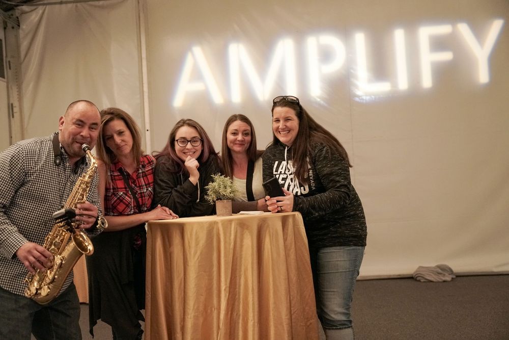 A group gathers joyfully beneath a glowing AMPLIFY neon sign, posing together at a table.