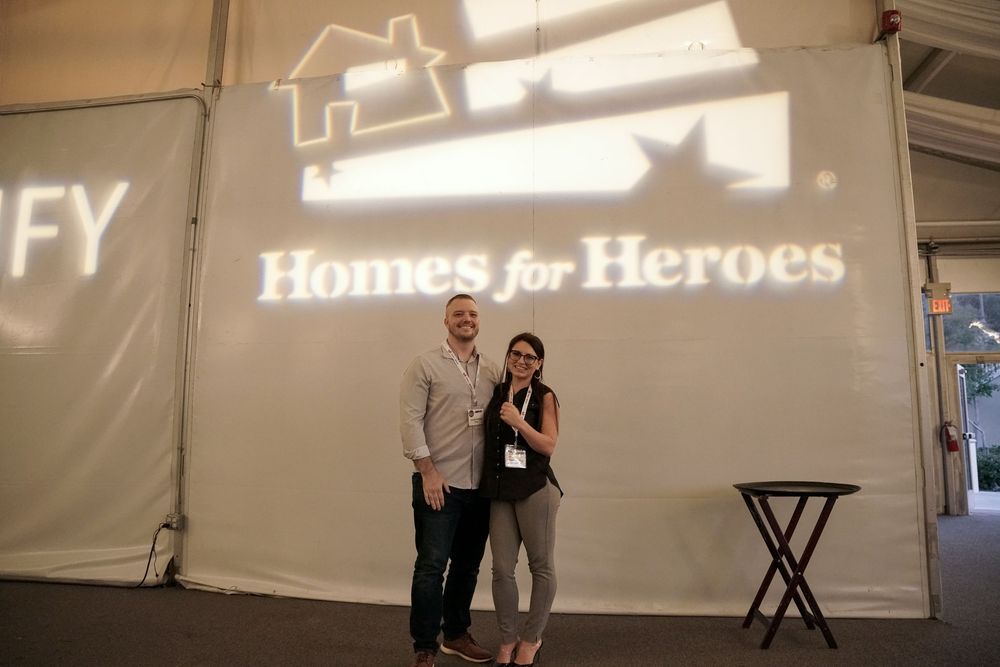 A couple stands together at a Home for Heroes event with illuminated signage behind them on a wall.