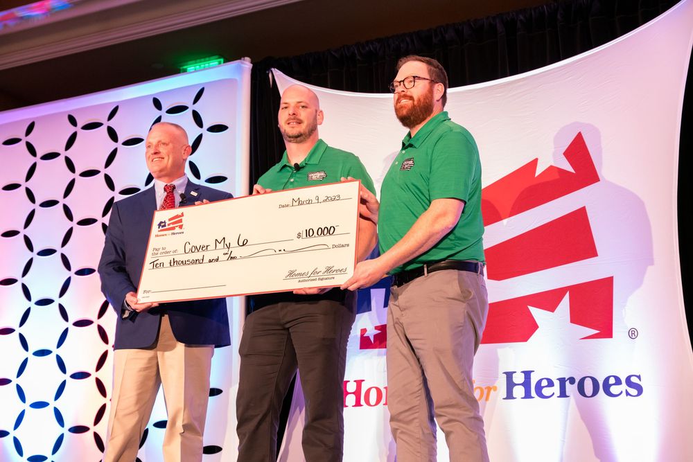 Three people stand on stage holding a large ceremonial check during an awards presentation with Heroes logo visible.