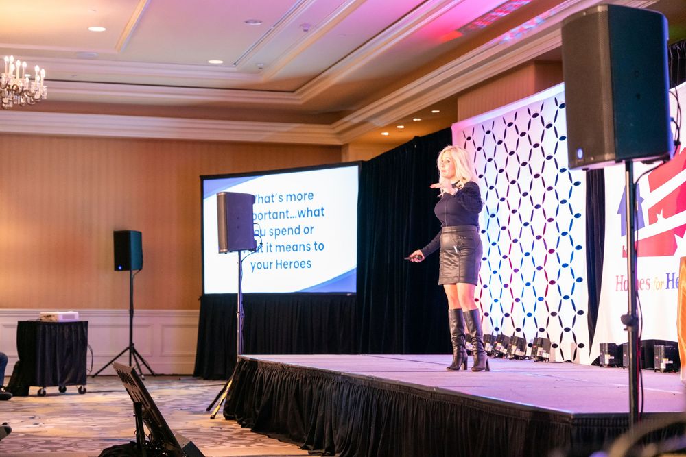Speaker presents on stage with a large projection screen and geometric pattern backdrop at a business conference.
