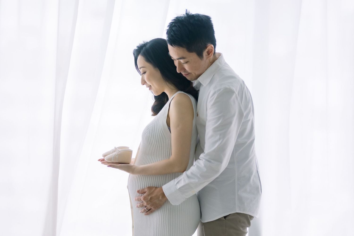 A tender maternity moment captured in elegant white, with soft window light illuminating an intimate moment between expectant parents.