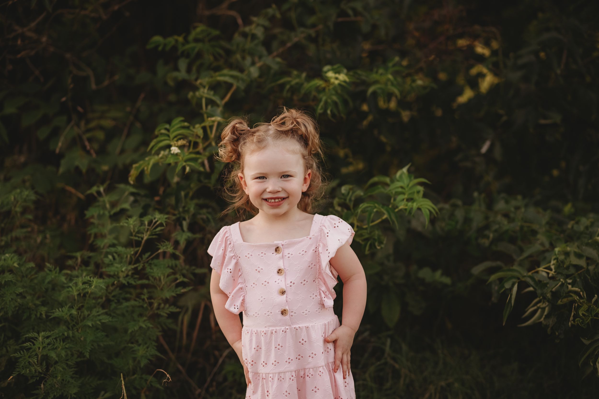 A pink-dressed toddler smiles while playing in a grassy field at sunset.
