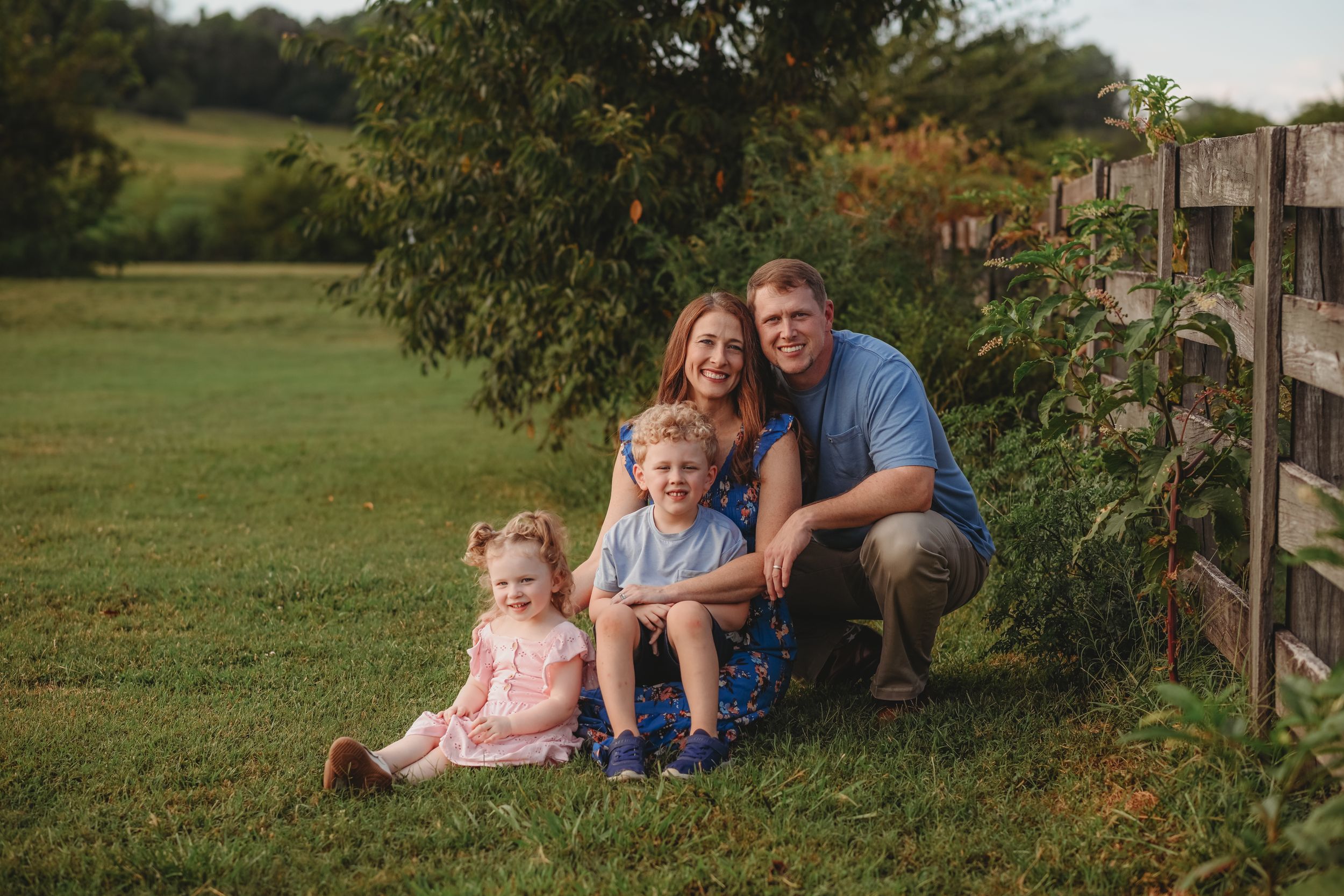 Family relaxes by a wooden fence with rolling farmland in the background.