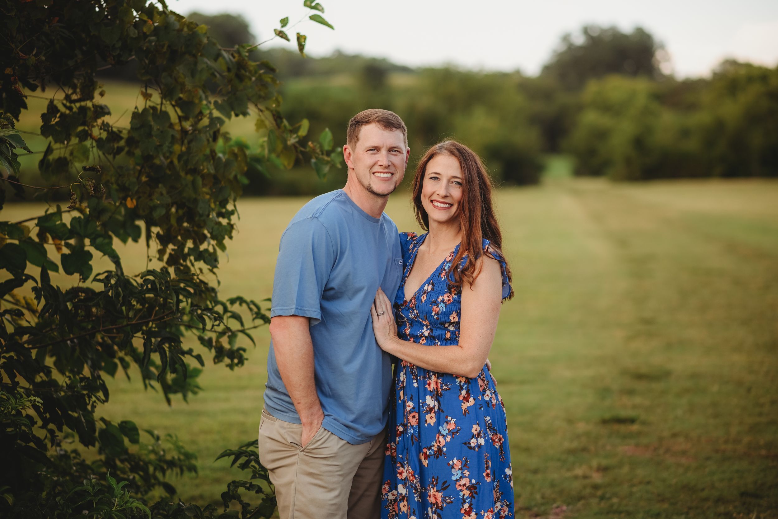 Couple poses together in casual attire against a scenic rural backdrop.