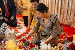 Traditional ceremony setup with person in grey dress arranging decorative items on red carpet floor.
