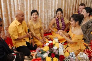 A group in yellow traditional attire celebrates at a festive gathering with flowers and drinks on the floor.